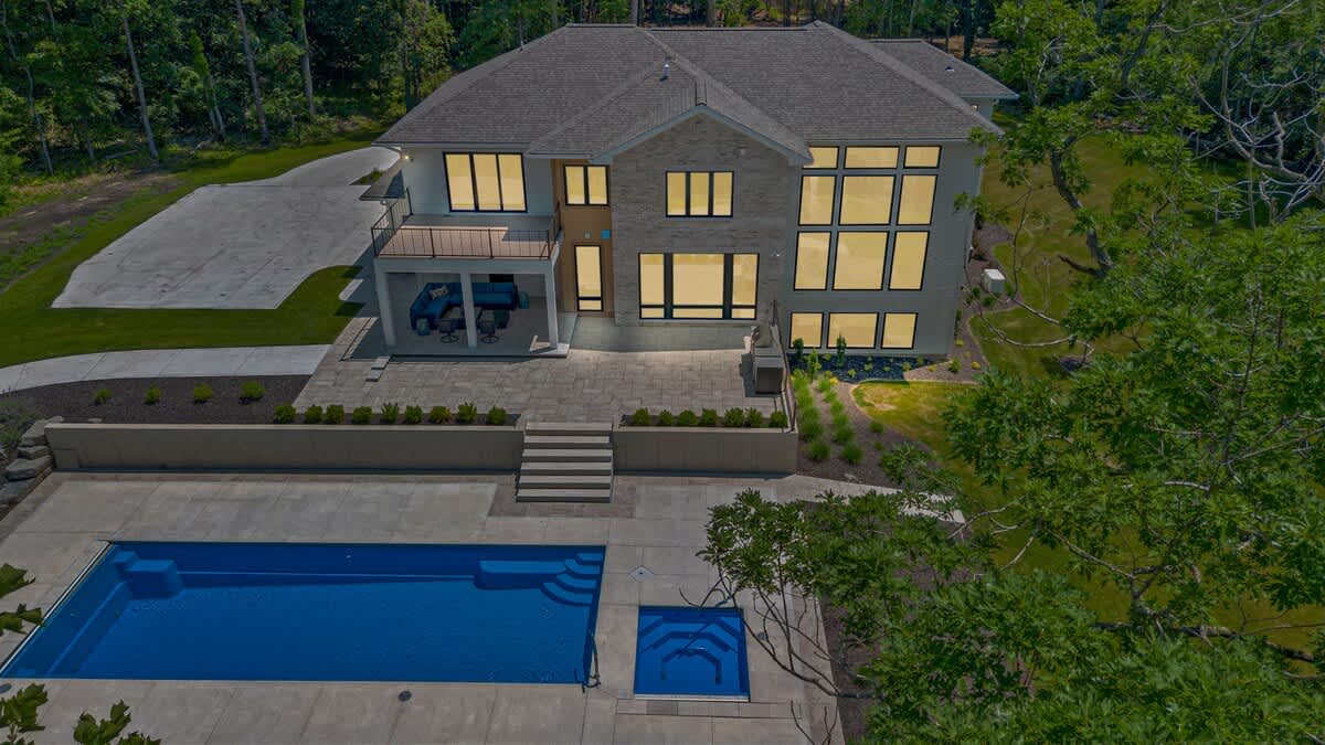 Rear exterior view of a large, modern house featuring extensive floor-to-ceiling windows, a covered outdoor dining area on a balcony, a spacious stone patio, and a rectangular swimming pool with an integrated hot tub.