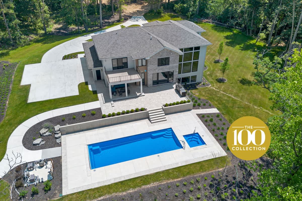 Aerial view of a modern house with a gray roof, light-colored exterior, and a large concrete driveway. A rectangular swimming pool with a smaller attached pool is visible in the foreground, surrounded by a patio and lush green landscaping.