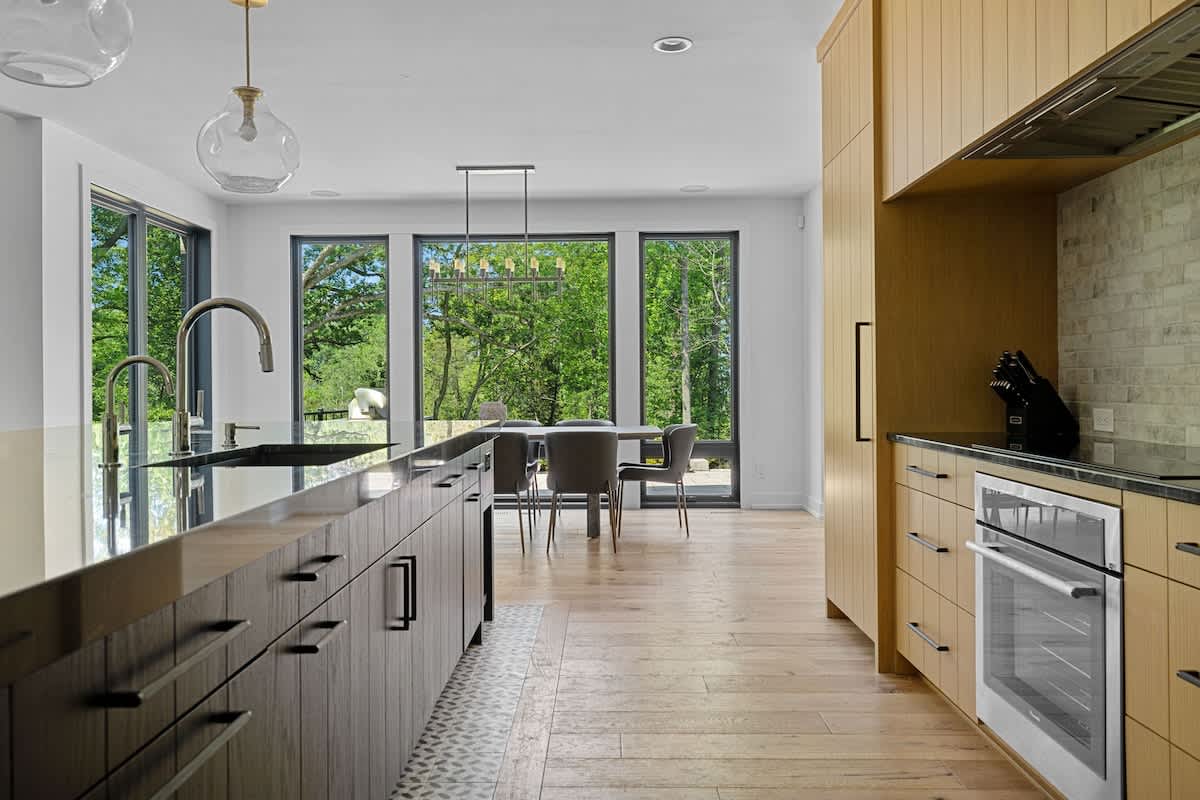 Modern kitchen with light wood cabinetry, a black island with a sink, and light wood floors. Beyond the island, a dining area with a dark table and chairs is visible through large windows offering views of trees.