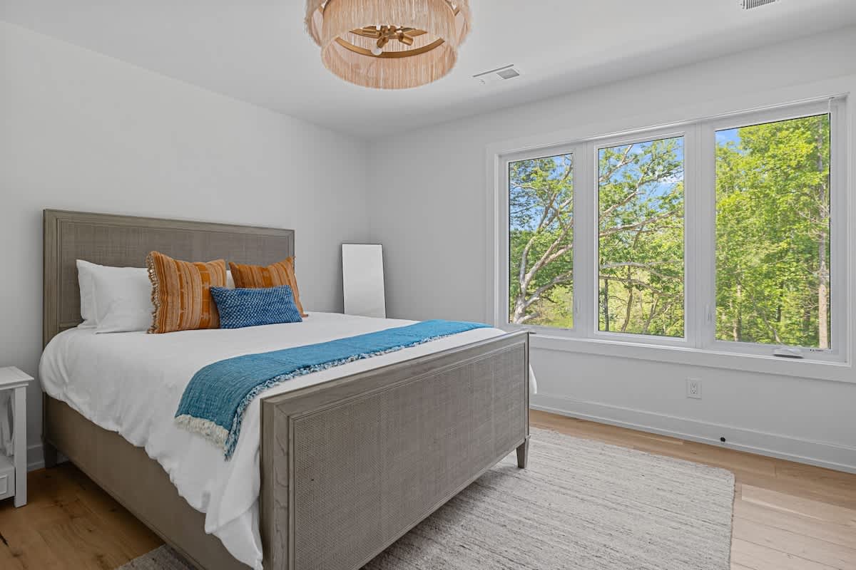 Bright bedroom-2 with a queen-sized bed, a light wood and woven headboard, a round woven chandelier, and large windows offering views of green trees.