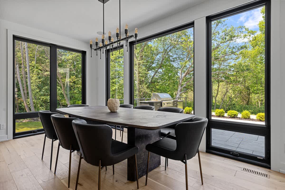 Elegant dining room with a long dark wooden table and eight dark chairs, surrounded by floor-to-ceiling windows offering views of a wooded backyard and an outdoor grill. A modern chandelier hangs above the table.