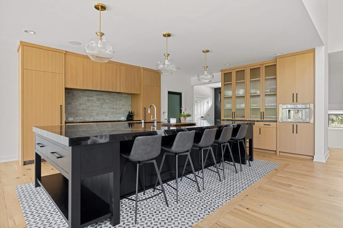 Modern kitchen featuring light wood cabinetry, a large black island with seating for five, and black countertops. Gold pendant lights hang above the island, and a patterned tile rug defines the space.