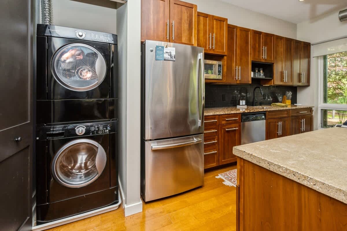 Compact laundry area with a stacked black washer and dryer unit, located adjacent to the kitchen with a stainless steel refrigerator and dark wood cabinets.