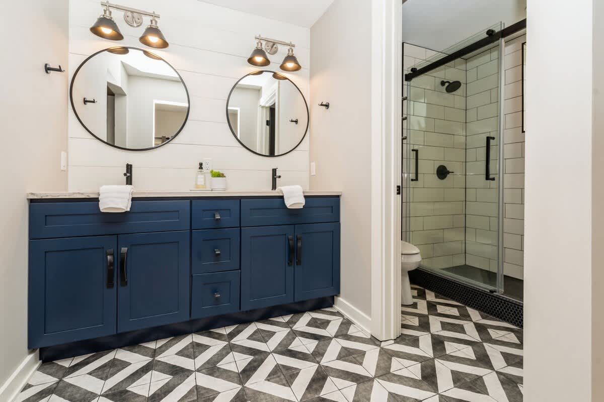 Stylish bathroom featuring a double vanity with navy cabinets, round mirrors, and a spacious walk-in shower with subway tiles and a black-framed glass door.