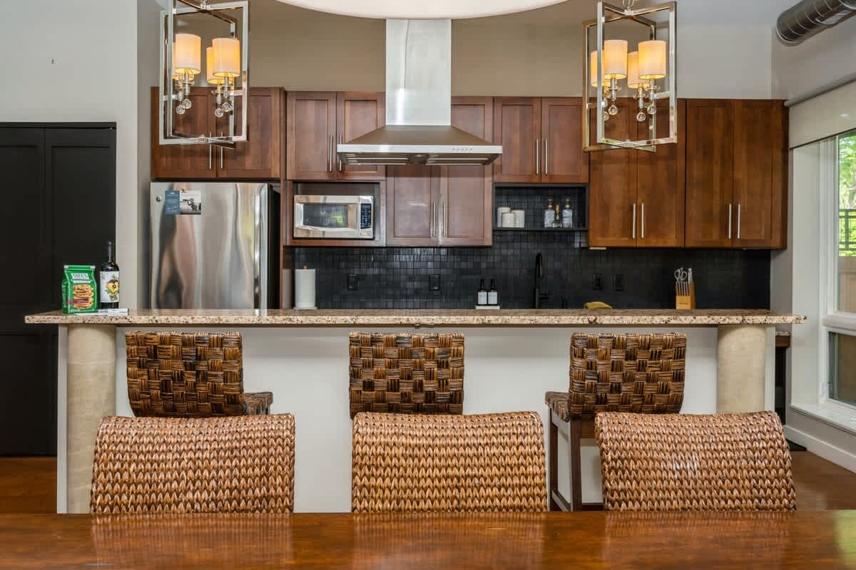 A detailed view of the kitchen island with four woven bar stools, showcasing the dark wood cabinetry, stainless steel appliances, and decorative pendant lighting above the counter.