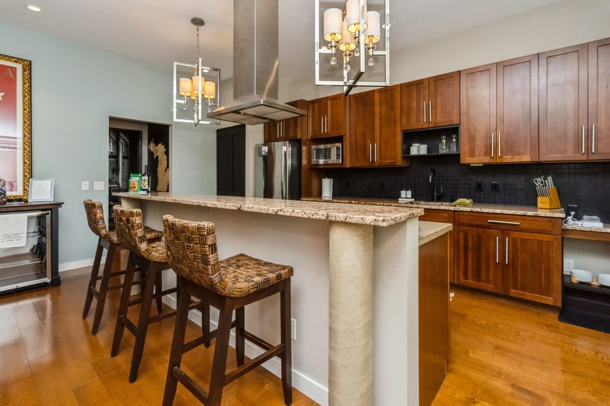 Modern kitchen featuring a large granite-topped island with three patterned bar stools, dark wood cabinetry, stainless steel appliances, and contemporary pendant lights.