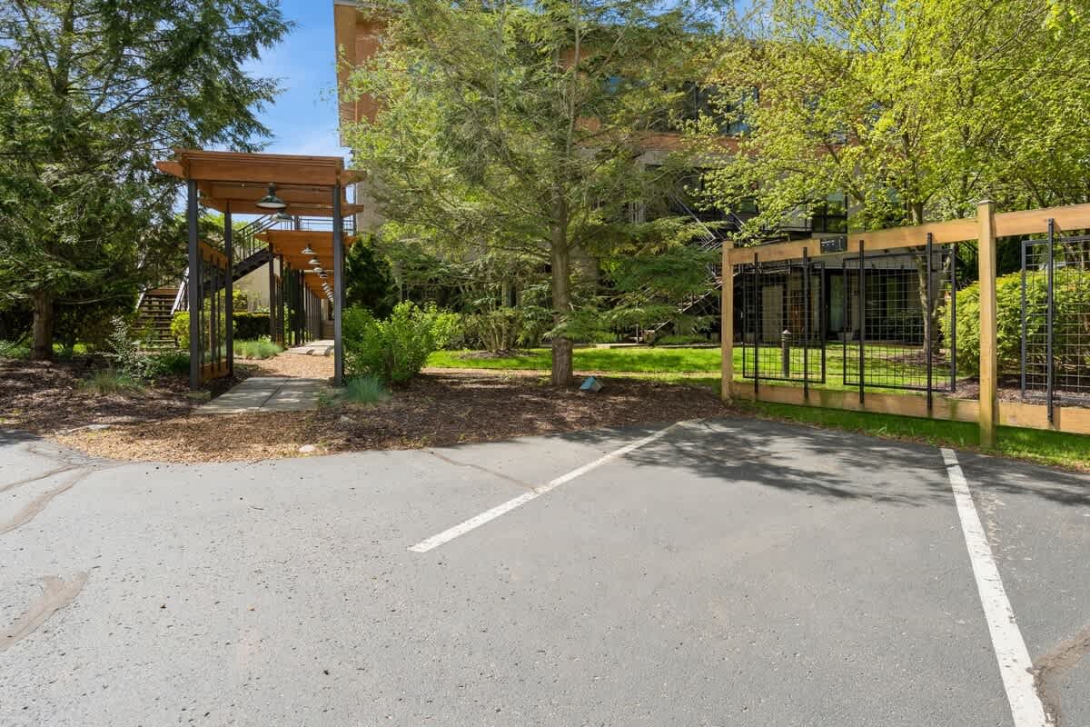 Front exterior view of the property entrance from a paved parking lot, featuring a gated walkway under a wooden pergola and mature trees.