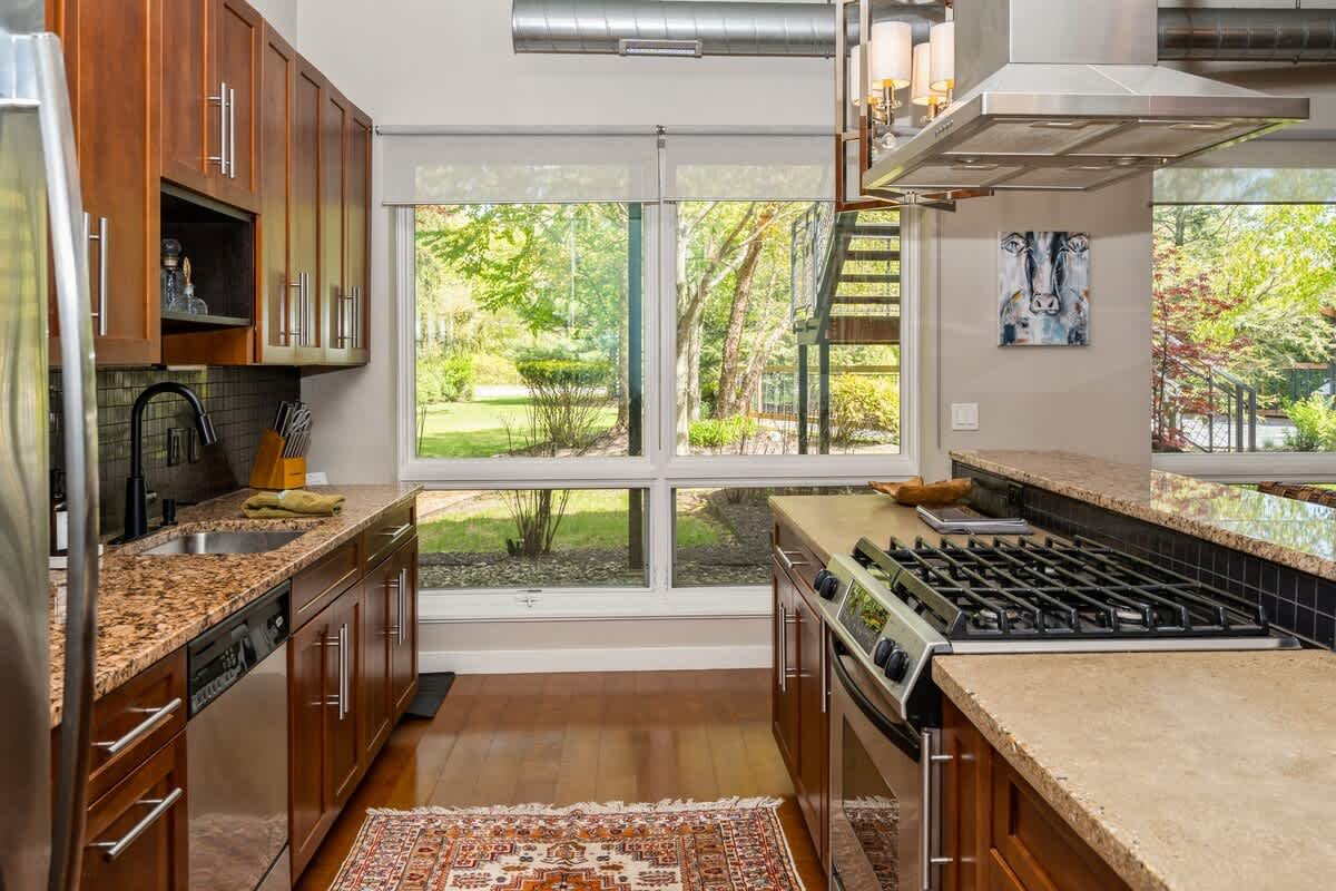 Bright kitchen with a gas range, granite countertops, dark wood cabinets, and a black tile backsplash, featuring large windows overlooking a lush green outdoor area.