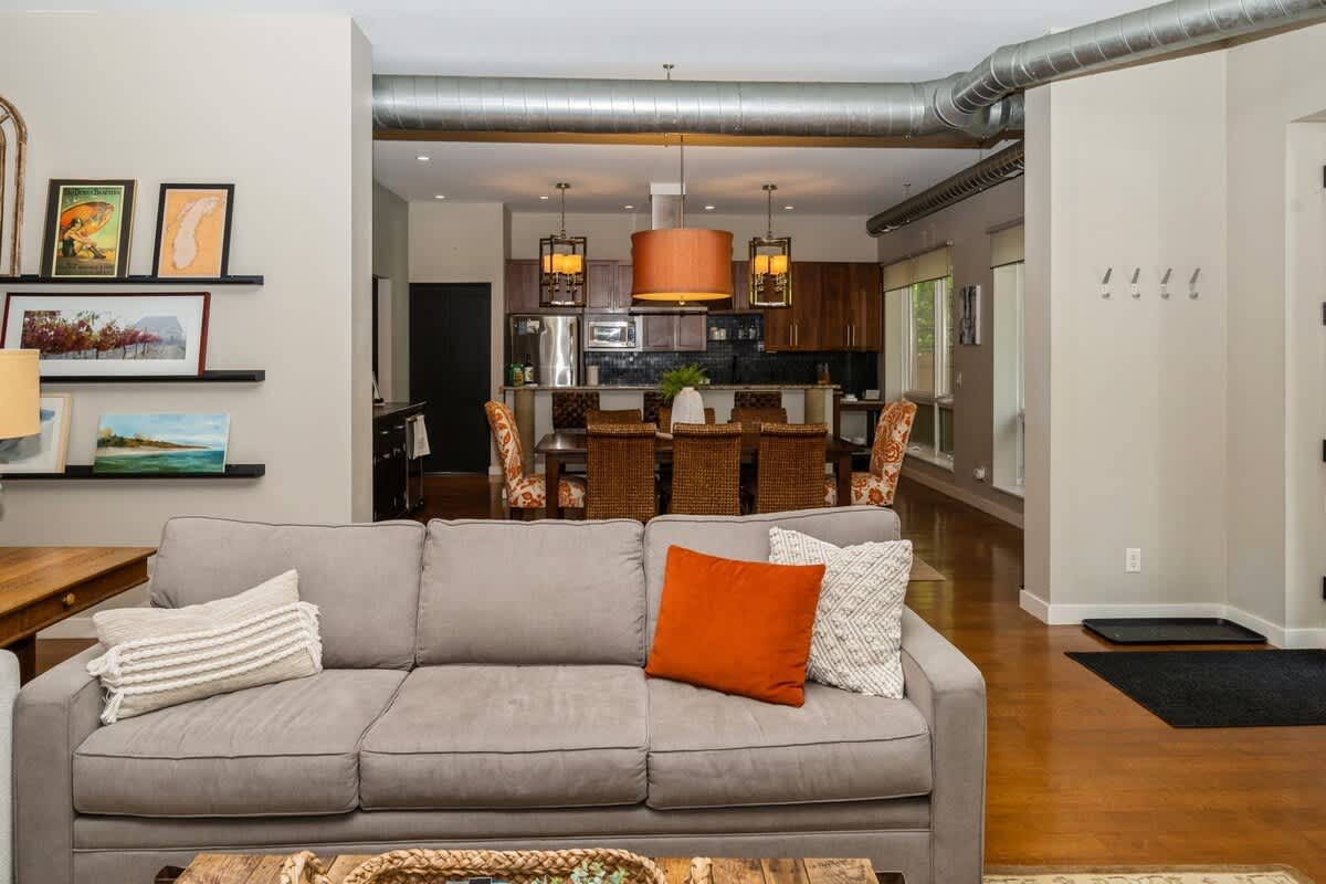 A wide view of the open-concept living area, featuring a large gray sofa and decorative wall shelves, with the dining area and kitchen visible in the background, connected by hardwood floors.