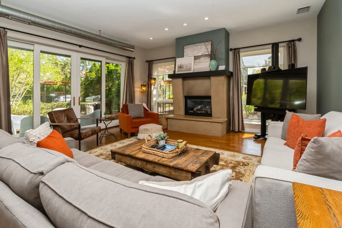 A spacious living room anchored by a large sectional sofa and an additional sofa, centered around a rustic wooden coffee table and a stone fireplace, with natural light from large glass doors.