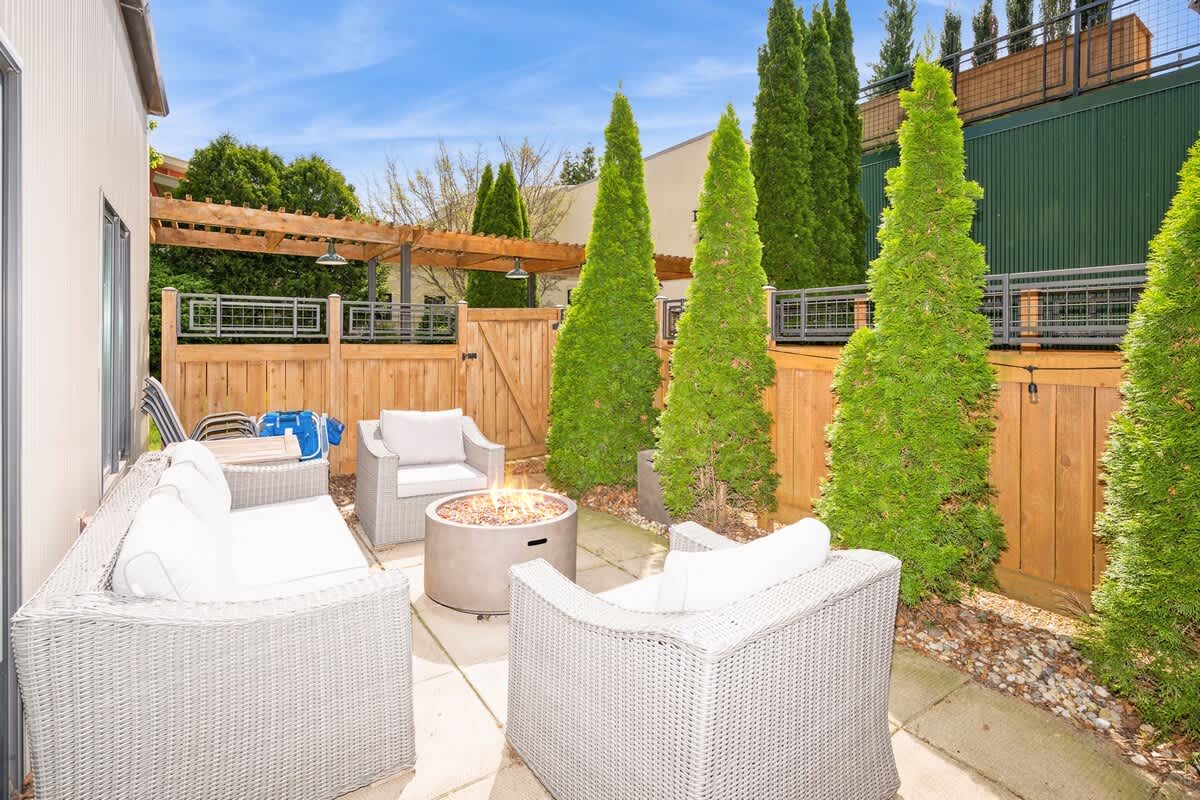 Inviting outdoor patio with light-colored wicker seating, a central fire pit, and tall evergreen trees providing privacy, enclosed by a wooden fence.
