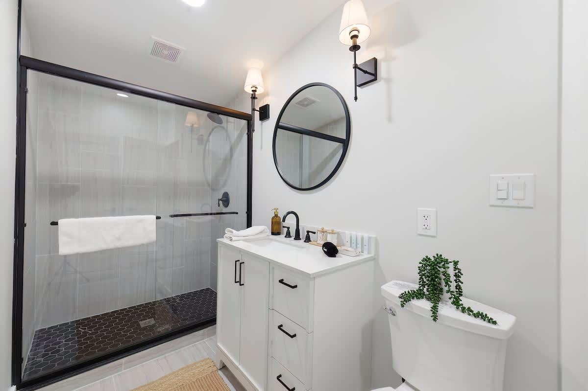 Modern bathroom featuring a white vanity with a black countertop and faucet, a round black-framed mirror, and a spacious walk-in shower with black hexagonal floor tiles and a glass enclosure.