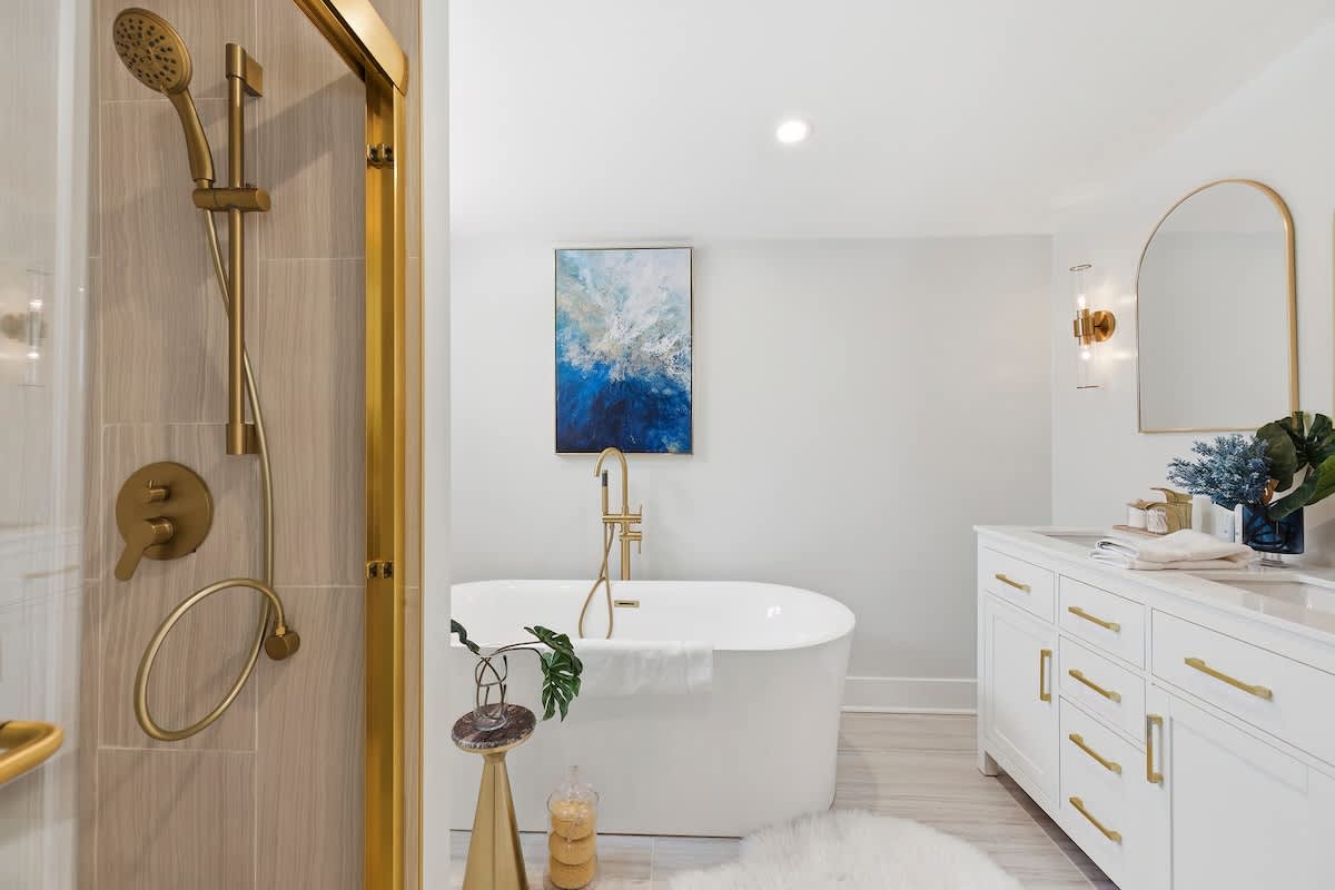 Modern bathroom featuring a freestanding white bathtub with a gold floor-mounted faucet, a double vanity with white cabinets and gold hardware, and a glass-enclosed shower with gold fixtures.