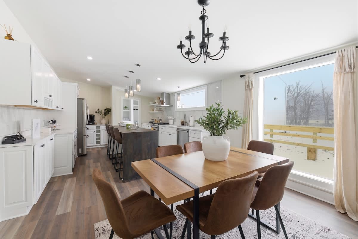 Open-concept dining area featuring a wooden dining table with black metal accents, surrounded by six brown leather chairs, and illuminated by a black chandelier, with a large window overlooking a snowy outdoor scene.
