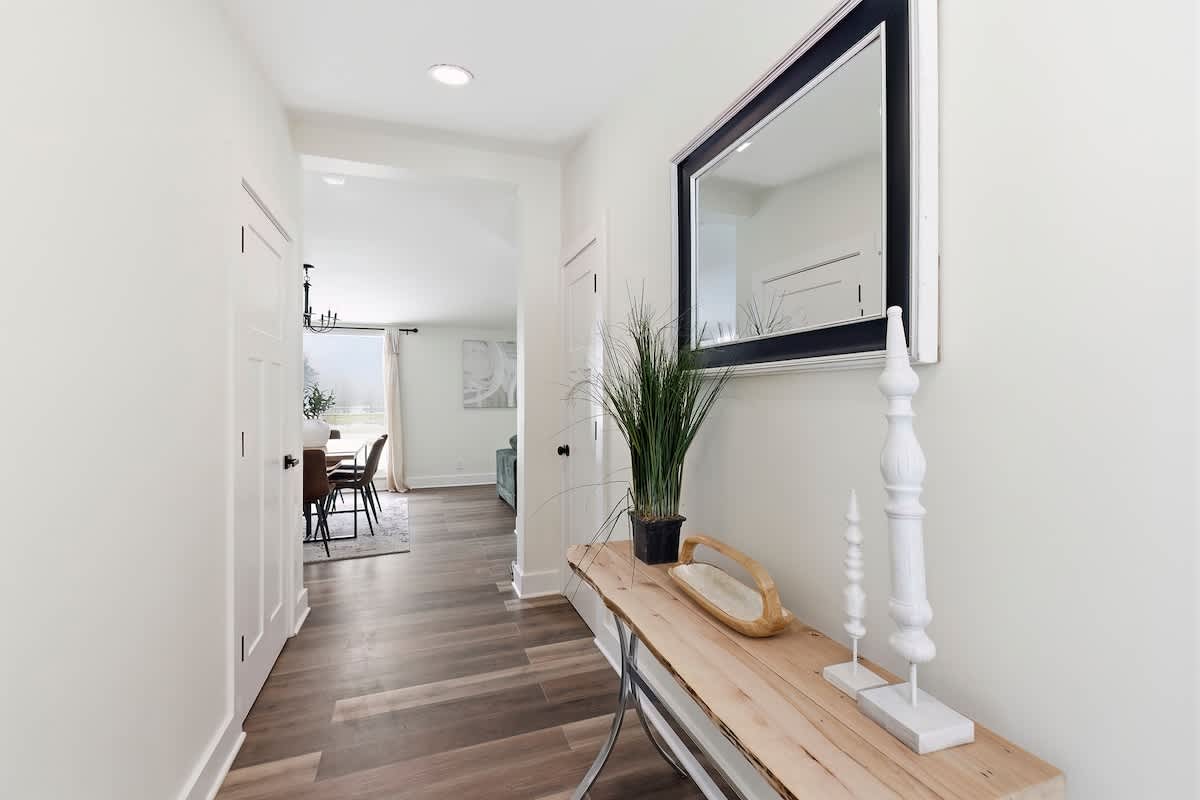 A bright and inviting foyer with light hardwood floors and white walls, featuring a rustic wooden console table adorned with decorative items and a large framed mirror. The hallway leads to other rooms in the house.