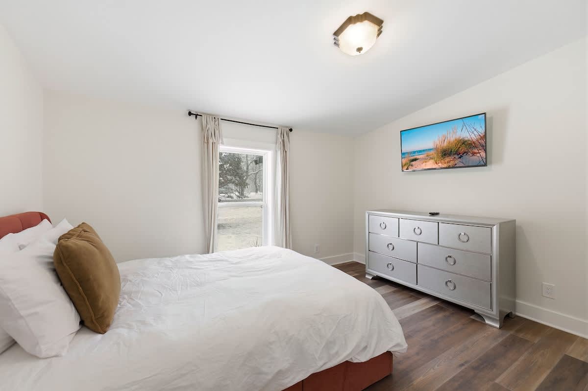 View of the same cozy bedroom, showing a large silver dresser with ring pulls, a wall-mounted TV, and a window with light-colored curtains overlooking a snowy outdoor scene.