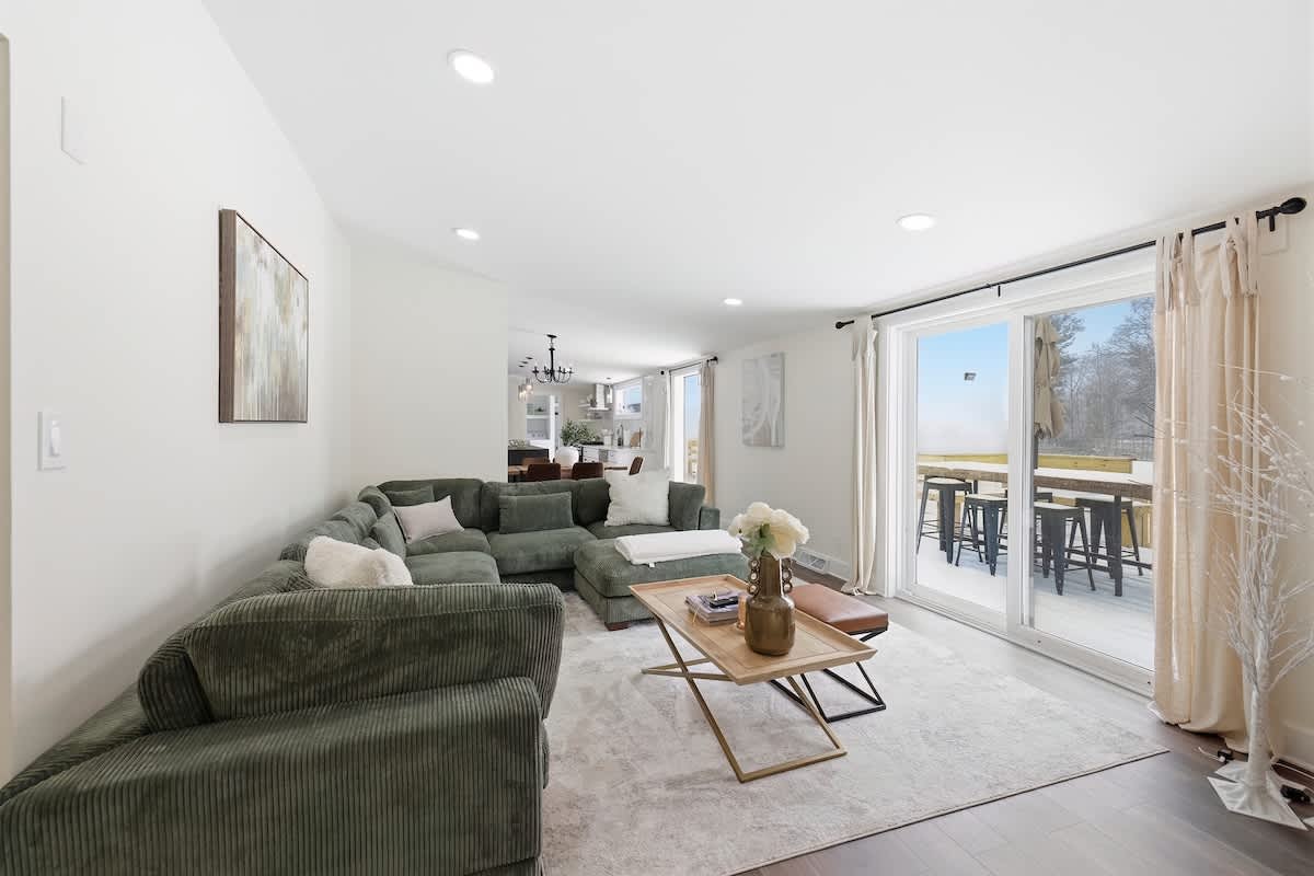 A spacious living room featuring a large green corduroy sectional sofa centered on a light area rug. Sliding glass doors open to an outdoor deck, and the room flows into the kitchen and dining area in the background.