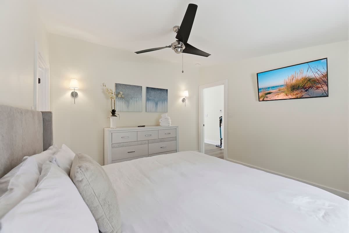 Bright bedroom with light cream walls, a large bed with white bedding, a black ceiling fan, a light grey dresser with modern art above it, and a wall-mounted TV displaying a beach scene.
