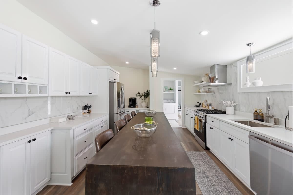 Wide view of a bright, modern kitchen featuring a long dark wood island, white shaker cabinets, stainless steel appliances including a refrigerator and gas range, and light wood-look flooring.
