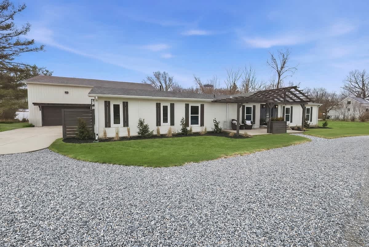 The front exterior of a white single-story house with dark shutters and a dark pergola over a patio area. A gravel driveway leads to the house, surrounded by green grass and some trees.