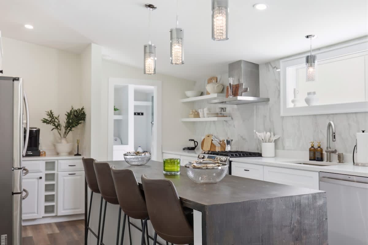 Modern kitchen with a long dark wood island featuring four brown leather bar stools, white cabinets, stainless steel appliances, and three pendant lights hanging above the island.