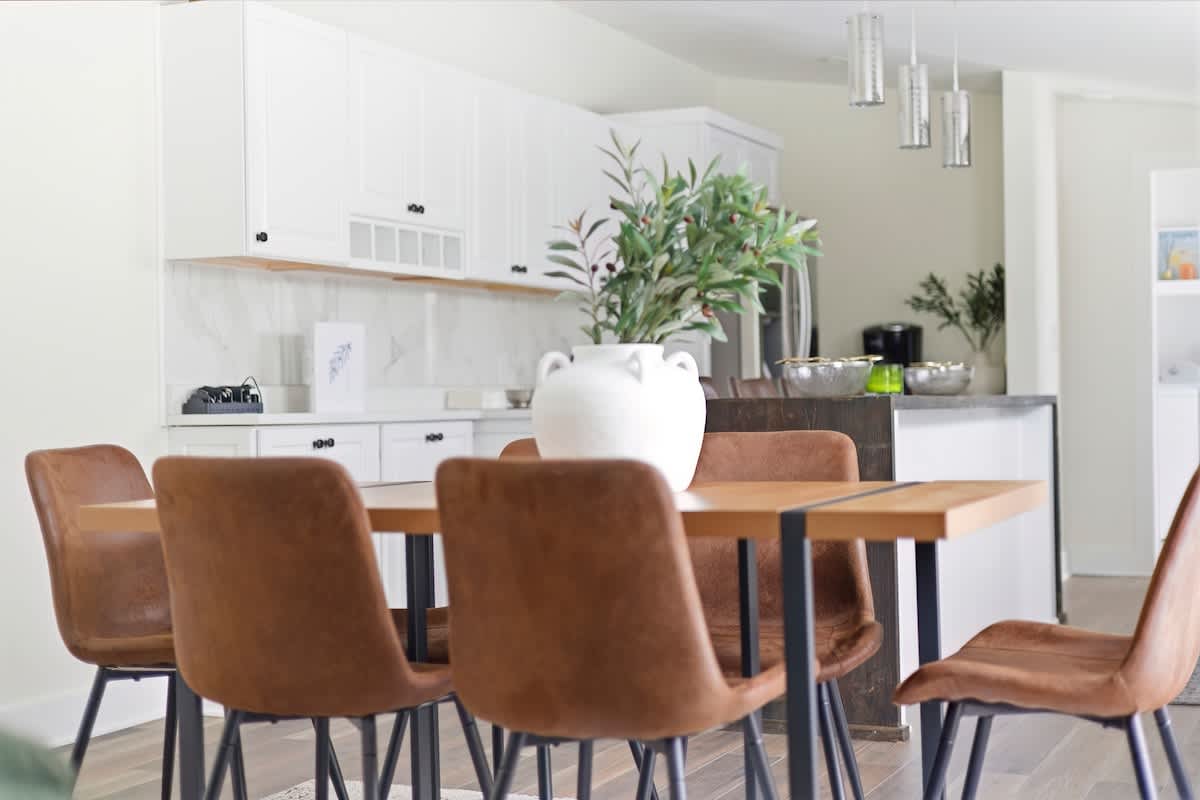 Close-up of the dining area showing a wooden dining table with black metal accents, surrounded by brown leather chairs, with a white vase holding greenery as a centerpiece, and a white kitchen with marble backsplash visible in the background.