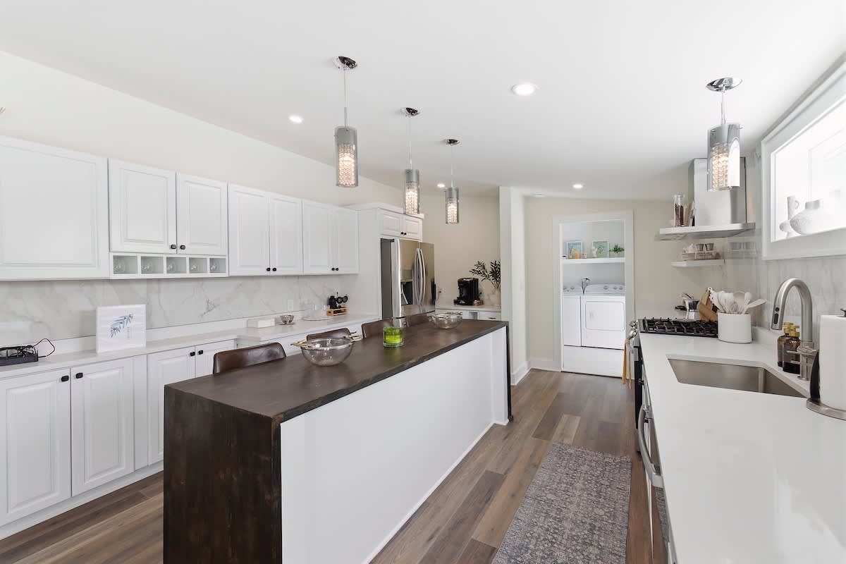 Spacious kitchen with white shaker cabinets, white countertops, a long dark wood island with seating, stainless steel appliances, and a view into a laundry area at the far end.