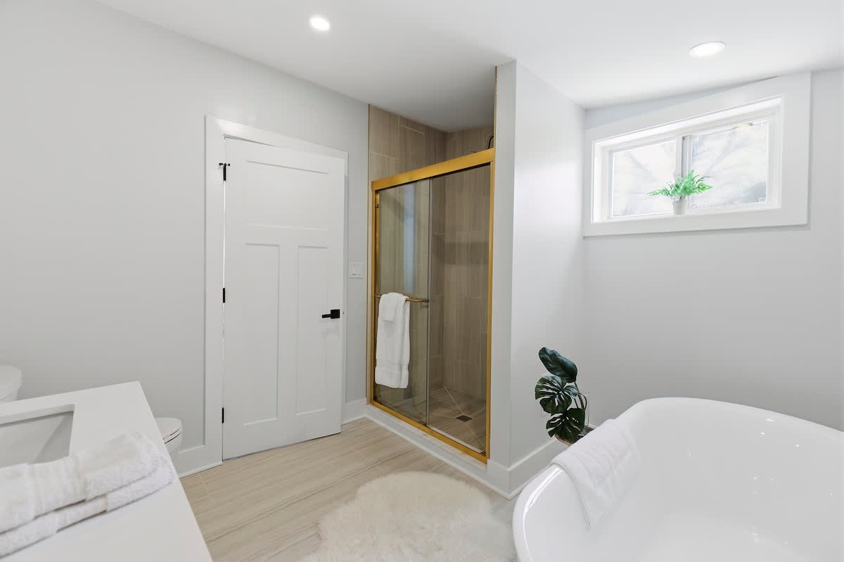 View of a modern bathroom showing a glass-enclosed shower with gold trim and a tiled interior, a freestanding white bathtub, and a white vanity, with light wood-look flooring.
