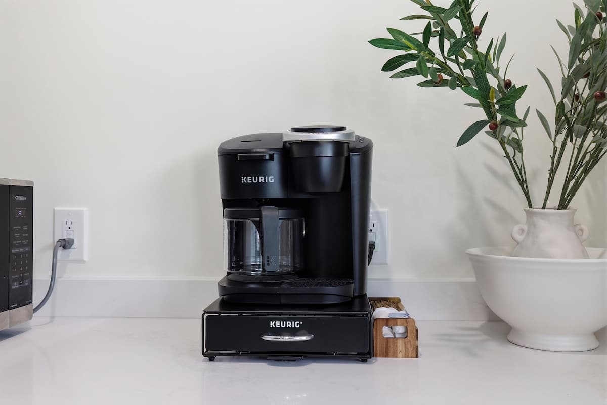 Close-up of a coffee station in the kitchen, featuring a black Keurig coffee maker on a black drawer organizer, next to a white vase with greenery, set against a light-colored wall.