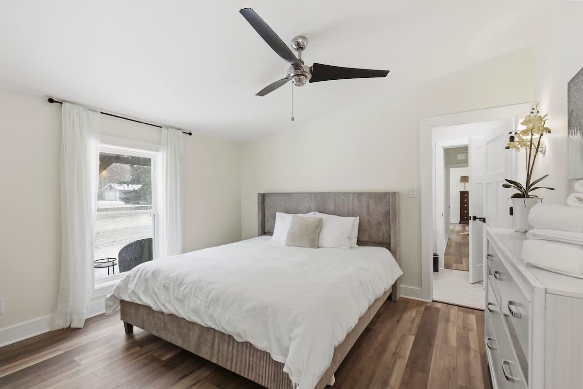 Cozy bedroom with a queen-sized bed featuring a gray upholstered headboard, a white dresser, and a ceiling fan, with a window offering an outdoor view.
