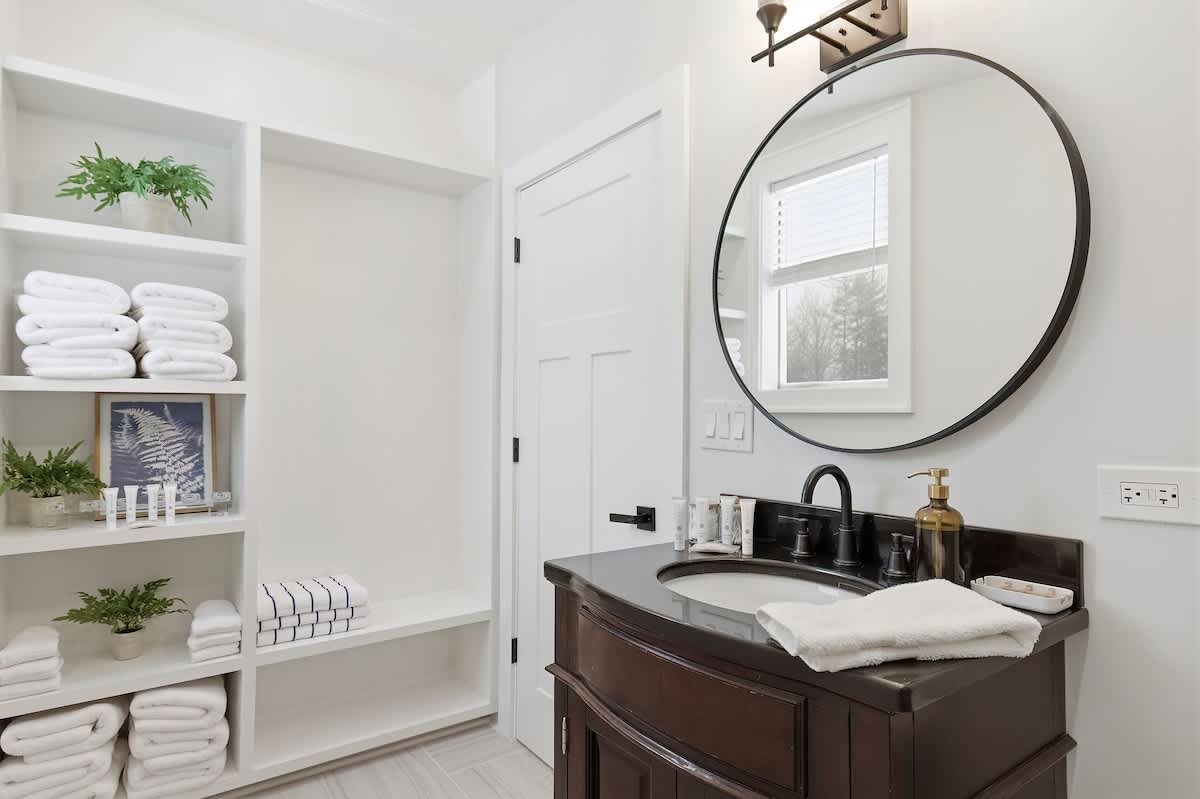 Another view of the modern bathroom, highlighting built-in white shelving stocked with neatly folded white towels and decorative plants, next to the dark wood vanity and round mirror.