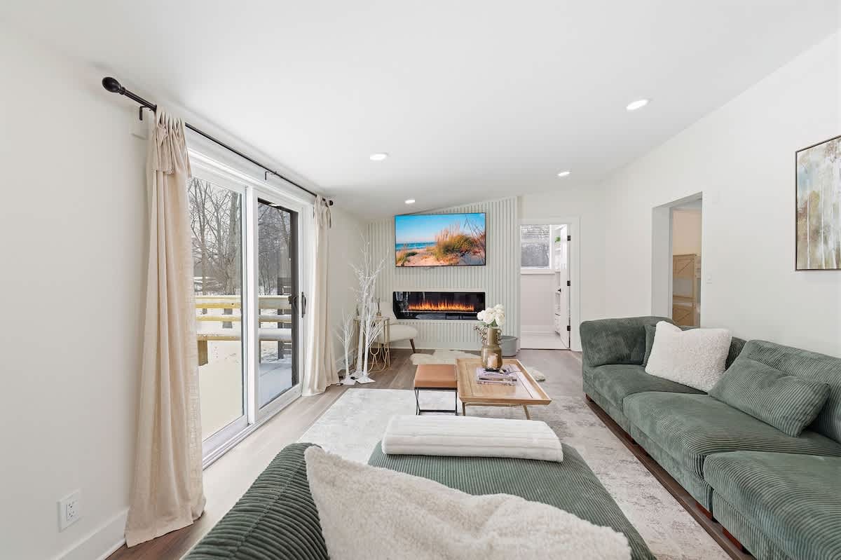 A cozy living room with a large green sectional sofa and a light-colored area rug. A wall-mounted TV is visible above a modern fireplace, with a sliding glass door leading to an outdoor area.