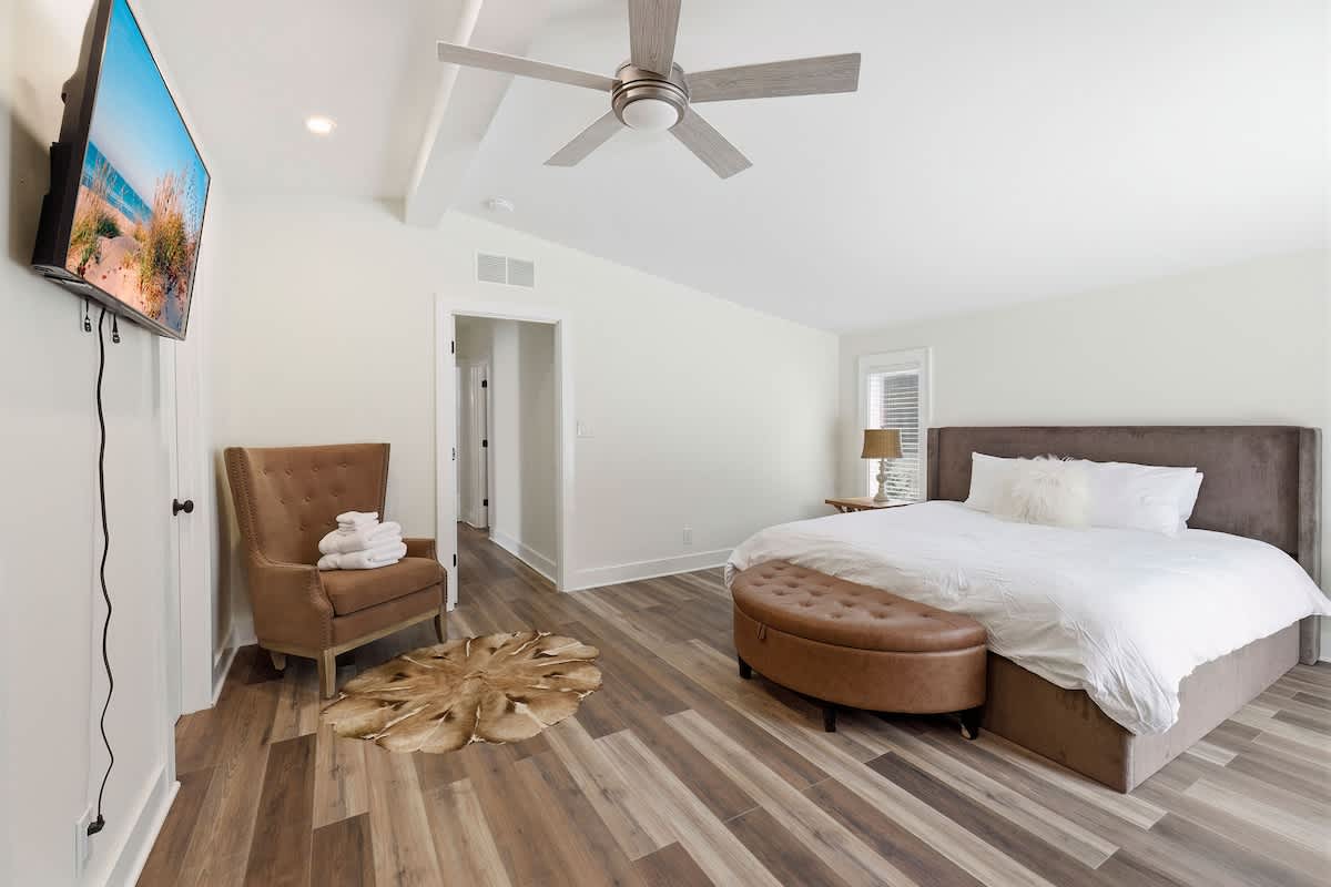 Bright bedroom with a large bed featuring a brown upholstered headboard, a brown leather ottoman at the foot, a brown armchair, and a wall-mounted TV, all on wood-look flooring.