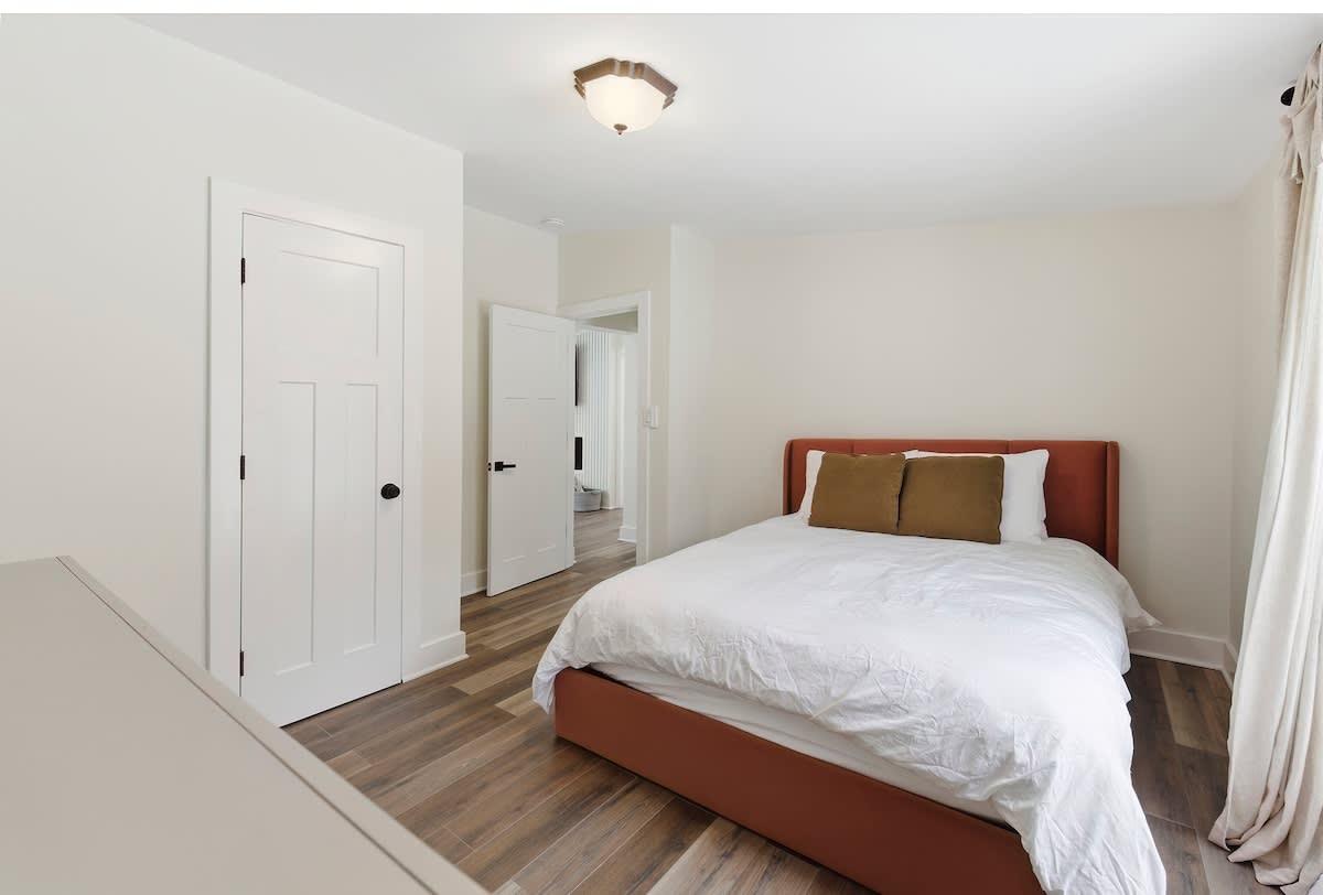Cozy bedroom with light walls, a queen-sized bed with an orange upholstered headboard and white bedding, and light wood-look flooring. A white dresser is partially visible on the left.