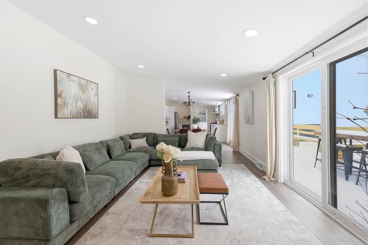 A wide view of the open-concept living room, showcasing a large green sectional sofa and a light area rug. Sliding glass doors provide access to an outdoor space, and the room connects to the kitchen and dining area beyond.