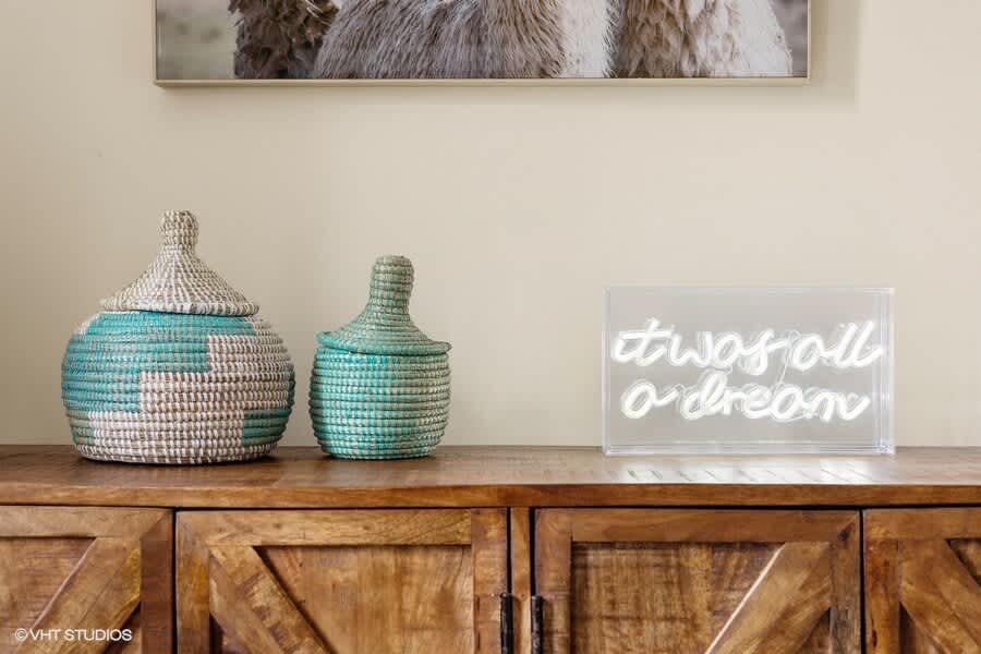 A rustic wooden console table featuring two woven lidded baskets in teal and natural tones, alongside a modern "it was all a dream" neon sign.