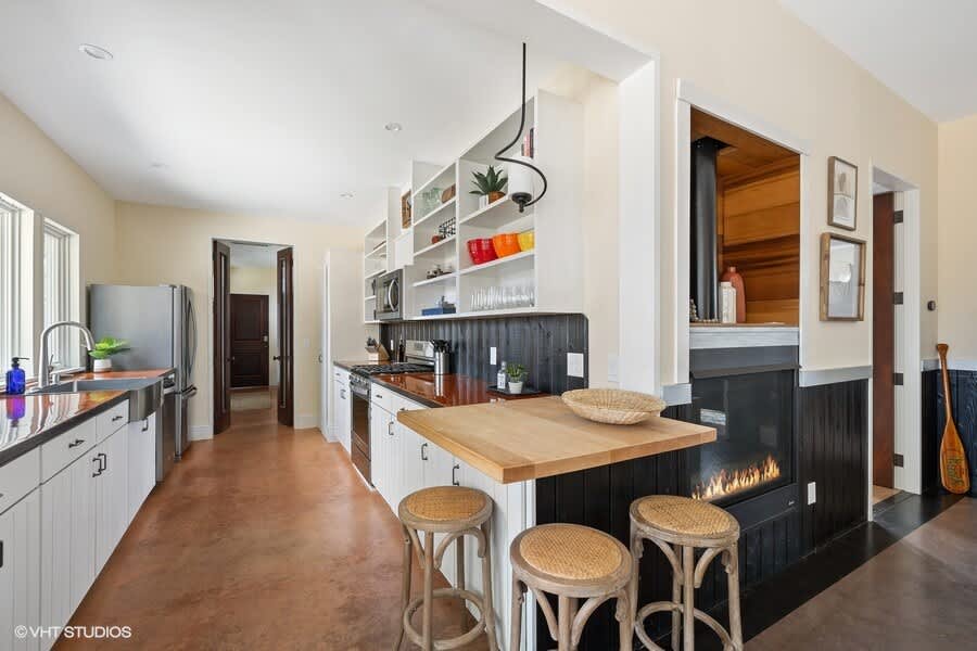 A modern kitchen with white cabinetry, dark countertops, open shelving, stainless steel appliances, and a breakfast bar with stools, featuring a built-in fireplace.