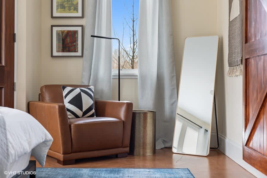 A cozy bedroom corner featuring a brown leather armchair with a geometric throw pillow, a modern black floor lamp, and a gold-framed full-length mirror.