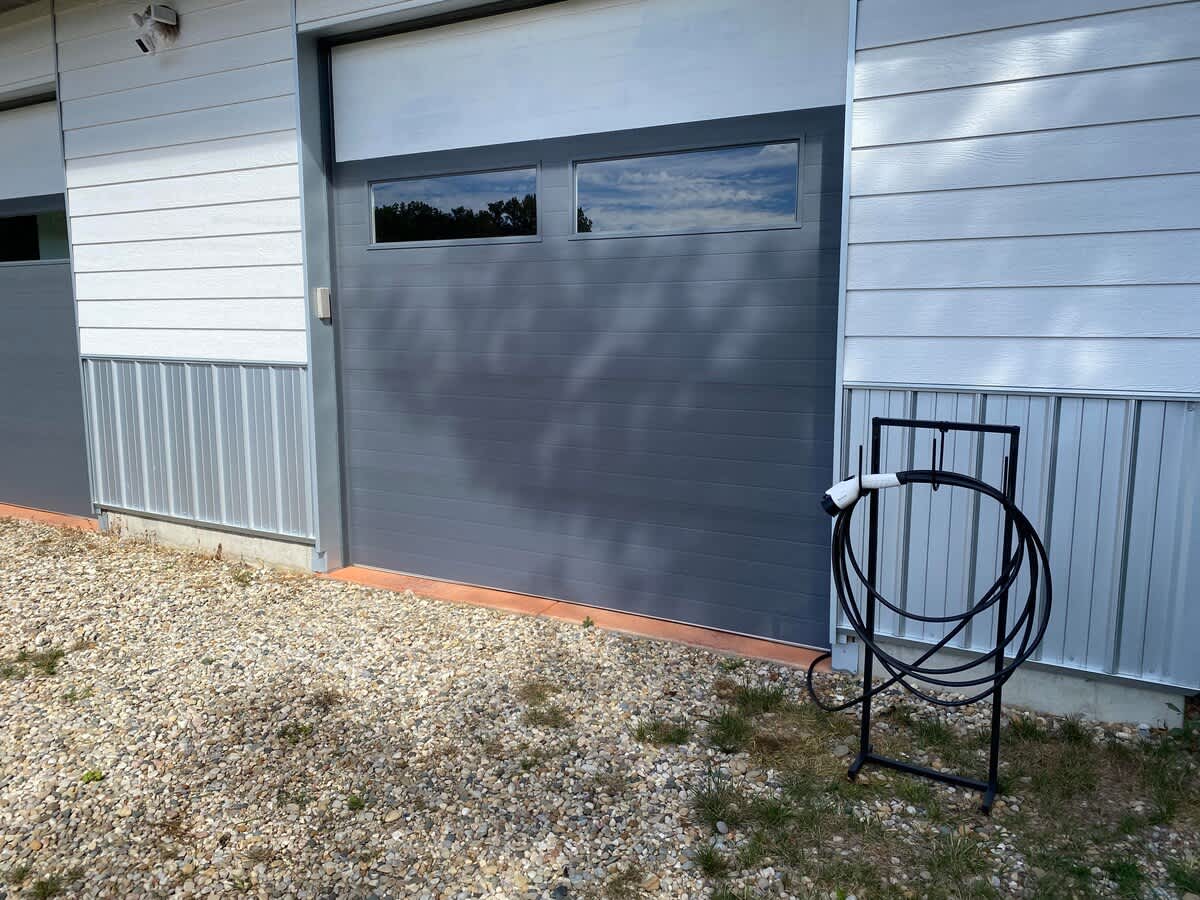 Modern gray garage door with two horizontal windows, flanked by white horizontal siding and corrugated metal panels, with an EV charging cable stand on a gravel and grass ground.