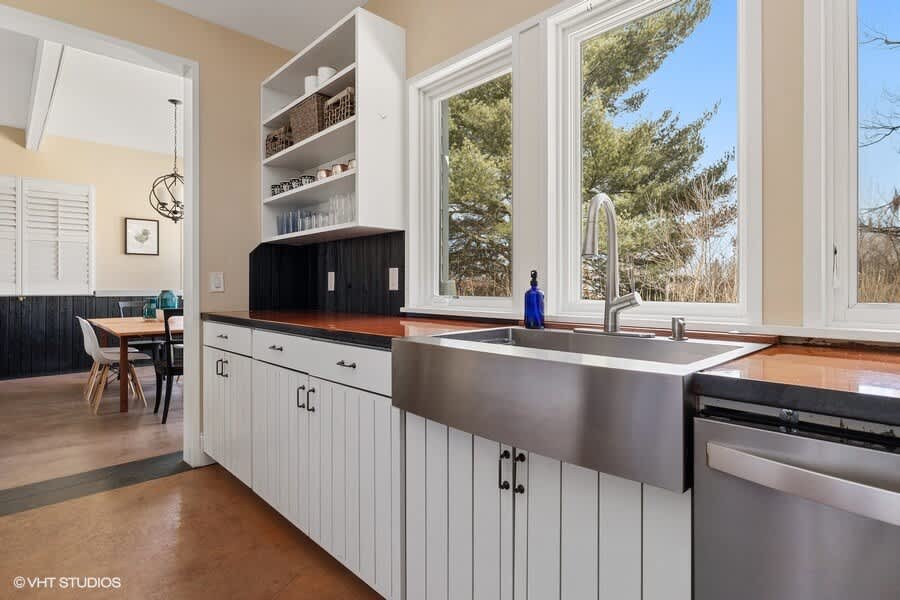 A bright kitchen corner with a large stainless steel farmhouse sink, dark countertops, white base cabinets, and expansive windows offering a view of the outdoors.