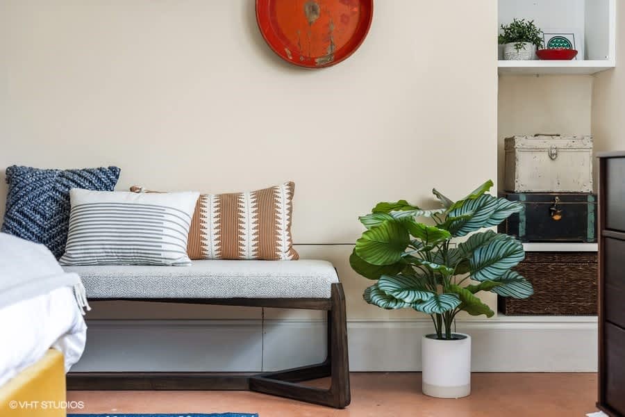 Detail shot of a bedroom corner featuring a light-colored bench with patterned pillows, a large potted plant, and built-in shelving with decorative items. The wall is a light cream color.