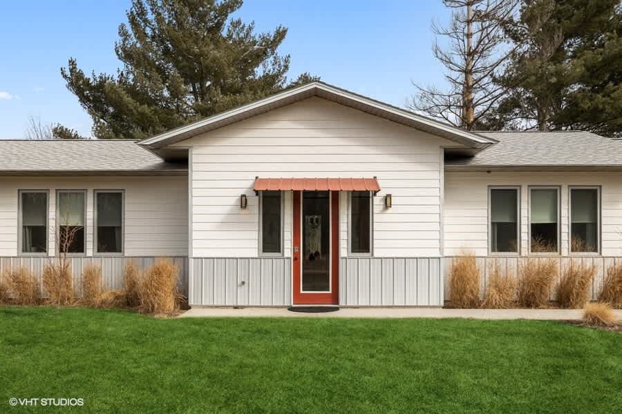 Modern single-story house with white horizontal siding and a lower band of corrugated metal, featuring a striking red front door beneath a matching red awning, flanked by windows and surrounded by green lawn and ornamental grasses.