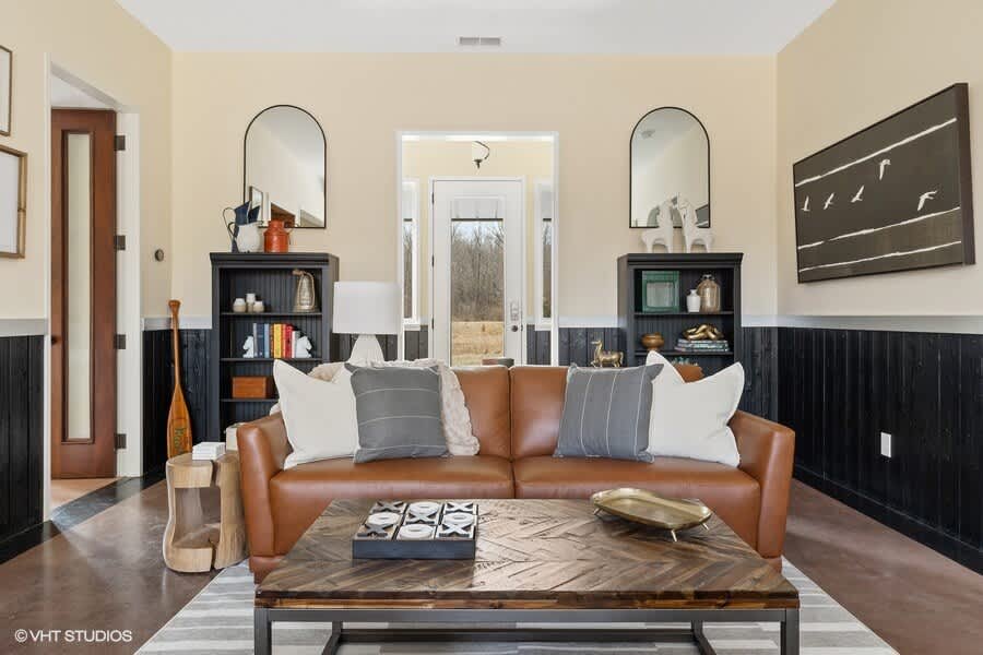 Cozy living room with a brown leather sofa, two black bookshelves flanking an entryway, and a wooden coffee table on a striped rug. Arched mirrors and artwork adorn the light yellow walls above dark wood paneling.