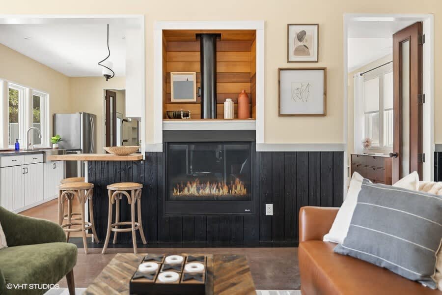 View of the living room centered around a modern fireplace with a black insert and dark wood paneling below, flanked by a kitchen bar area with stools and a brown leather sofa. Artwork hangs on the light yellow walls.