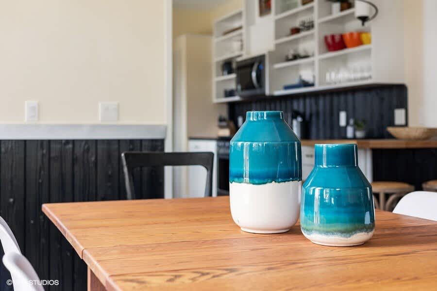 A close-up of a wooden dining table adorned with two teal and white ceramic vases, with the kitchen's open shelving and dark wainscoting visible in the background.
