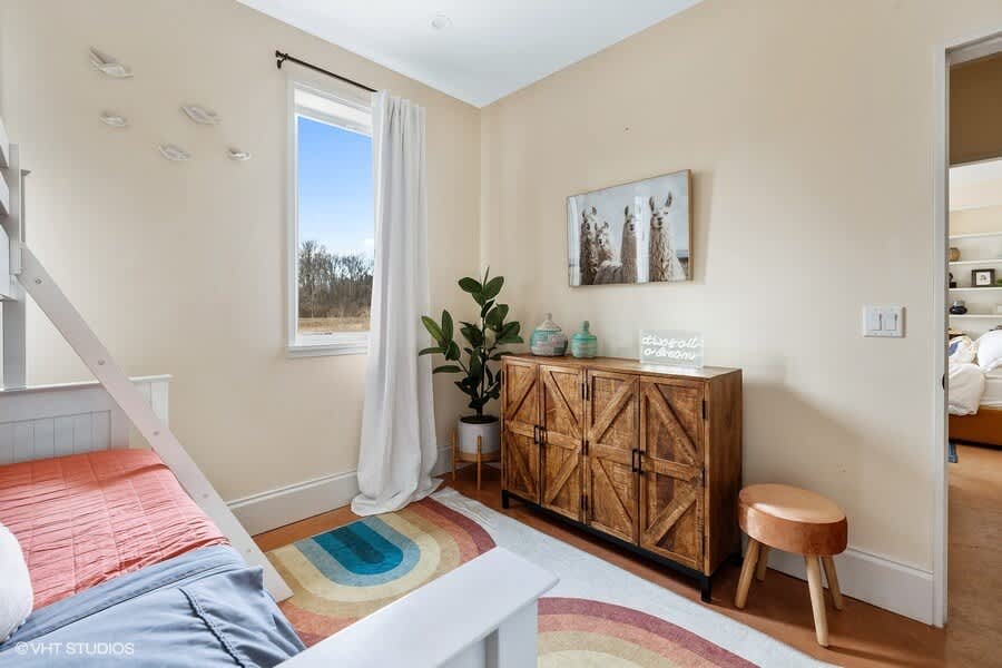 View of the kids' bedroom showing the white bunk beds, a rustic wooden cabinet, and a window with white curtains. A llama-themed picture hangs above the cabinet.