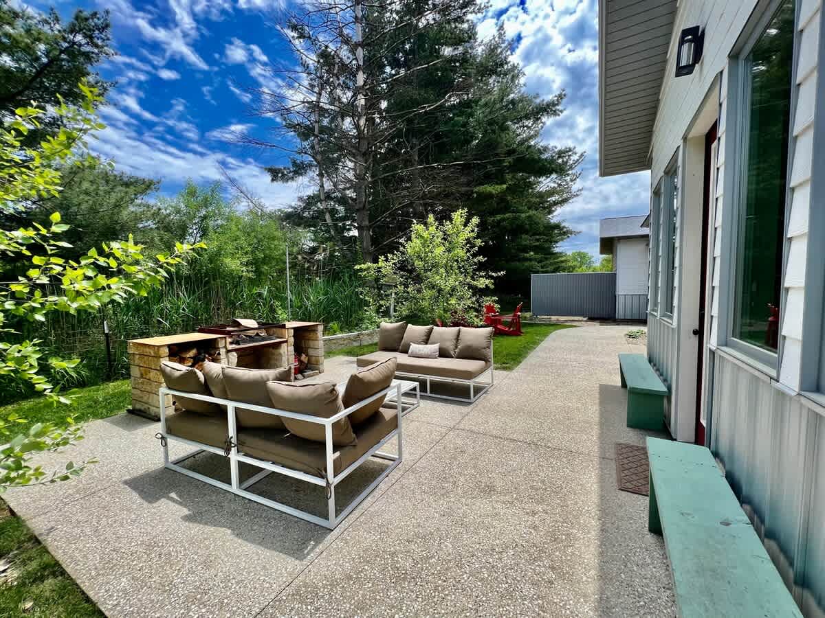 Expansive outdoor patio with modern seating, a built-in grill, and a view of the house exterior. The area is surrounded by green trees and natural landscaping.