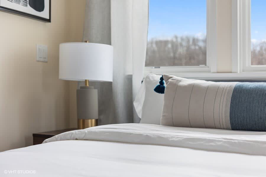 A close-up of the bed in a bedroom, showing white bedding, a blue and white decorative pillow with a tassel, and a modern table lamp on a nightstand.