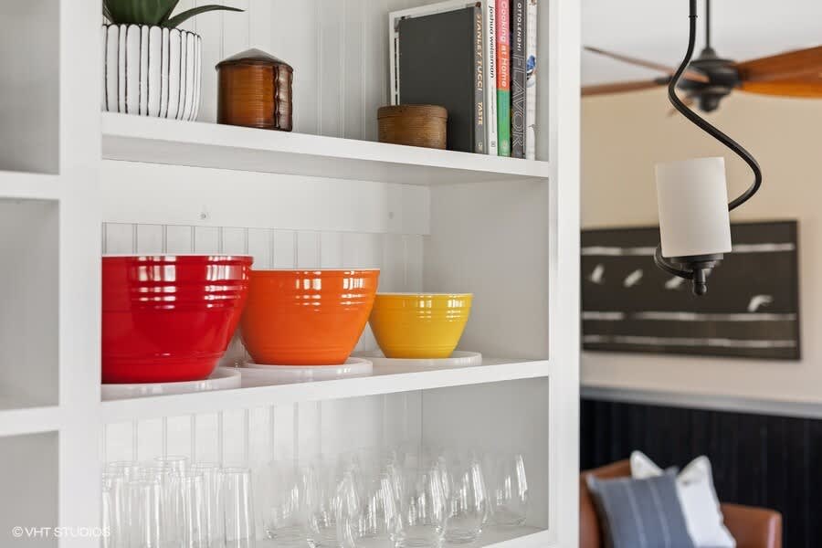 A detail shot of white open shelving displaying colorful red, orange, and yellow mixing bowls, clear drinking glasses, and various decorative items.