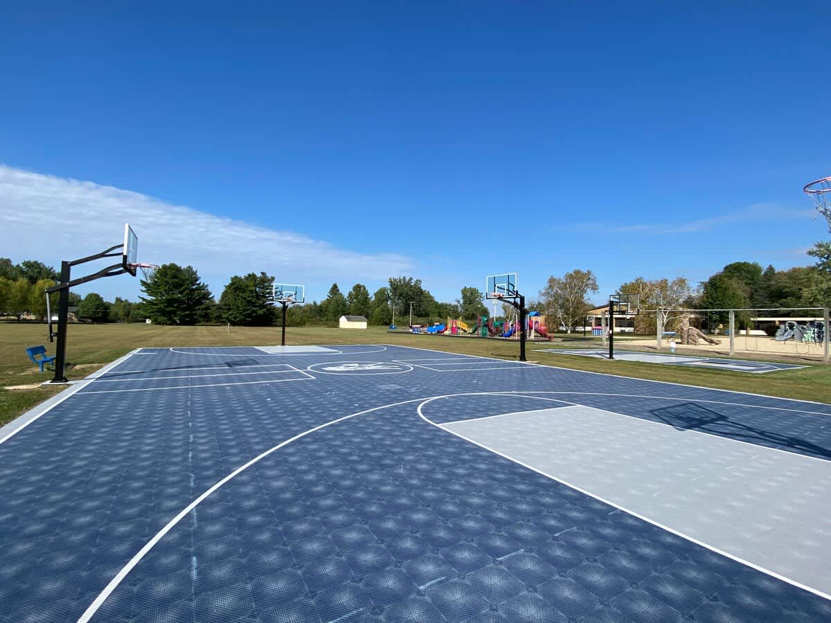 Expansive outdoor basketball court featuring blue and light gray interlocking tiles, multiple basketball hoops, and crisp white court lines, surrounded by a vibrant green lawn under a clear blue sky.
